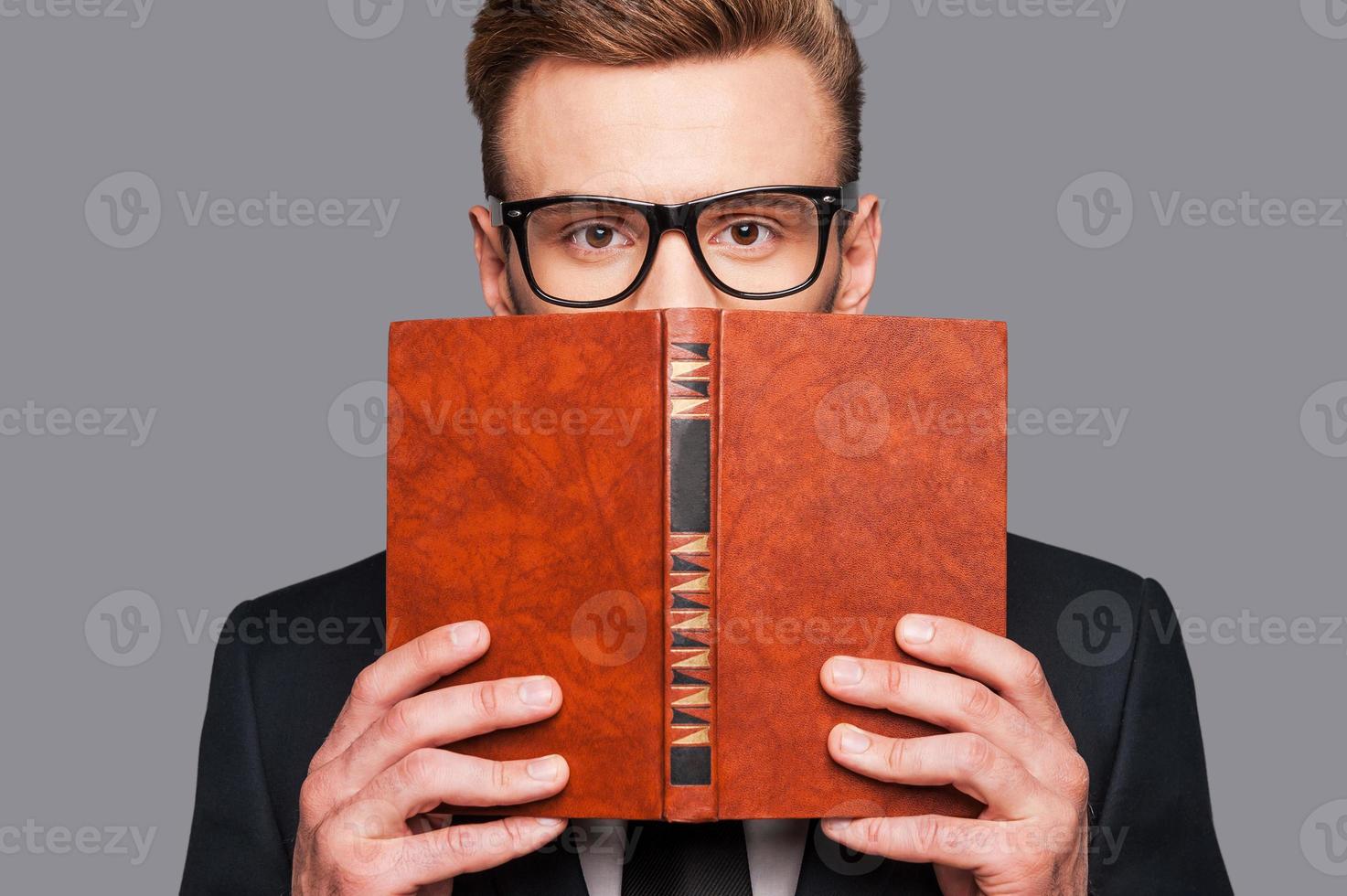 más conocimiento joven en ropa formal escondiendo su rostro detrás de un libro mientras está de pie contra el fondo gris foto