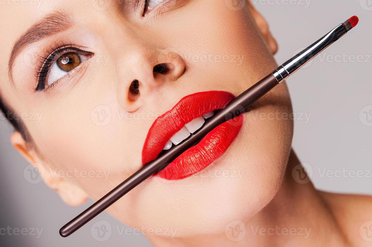 Cosmetics artistry. Close-up of a beautiful woman with red lips holding make-up brush in her mouth and looking away while standing against grey background photo