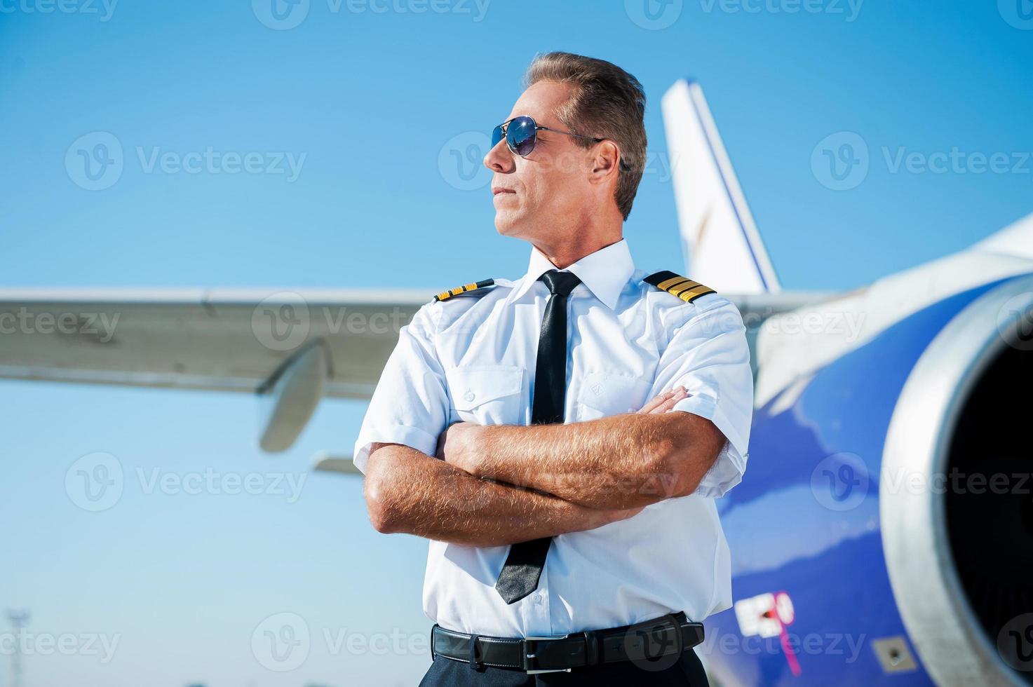 Passionate by sky. Confident male pilot in uniform keeping arms crossed and looking away with airplane in the background photo