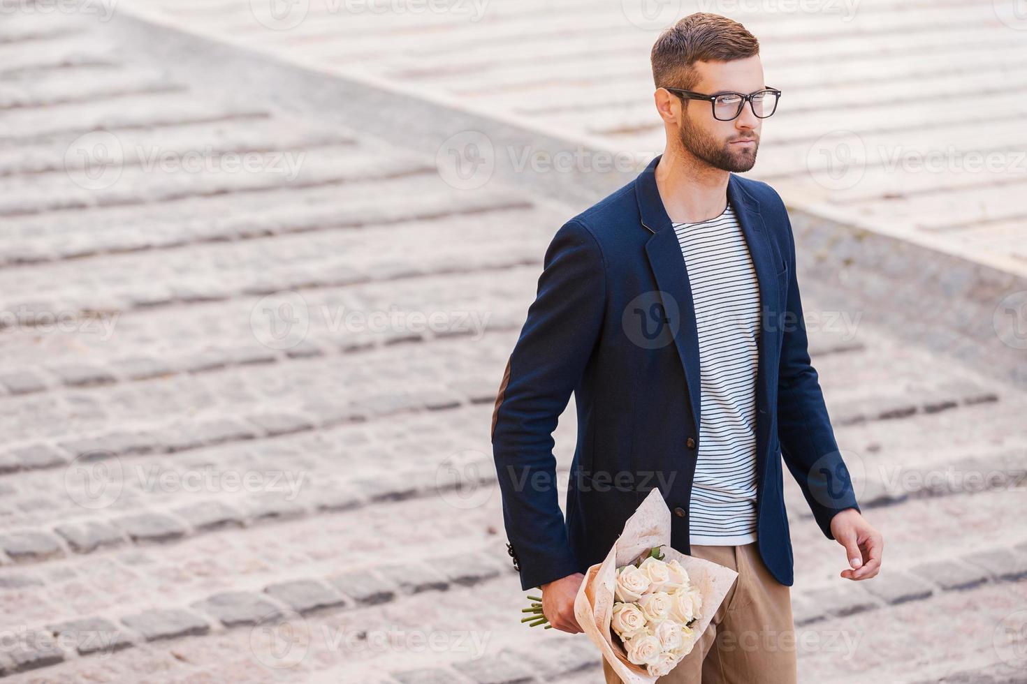 Hurrying to be in time. Handsome young man in smart casual wear holding bouquet of flowers while walking by the street photo