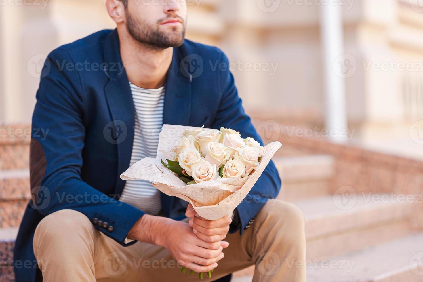 esperando a su novia. primer plano de un joven apuesto con una chaqueta inteligente sosteniendo un ramo de flores y sentado en la escalera cerca de la casa foto