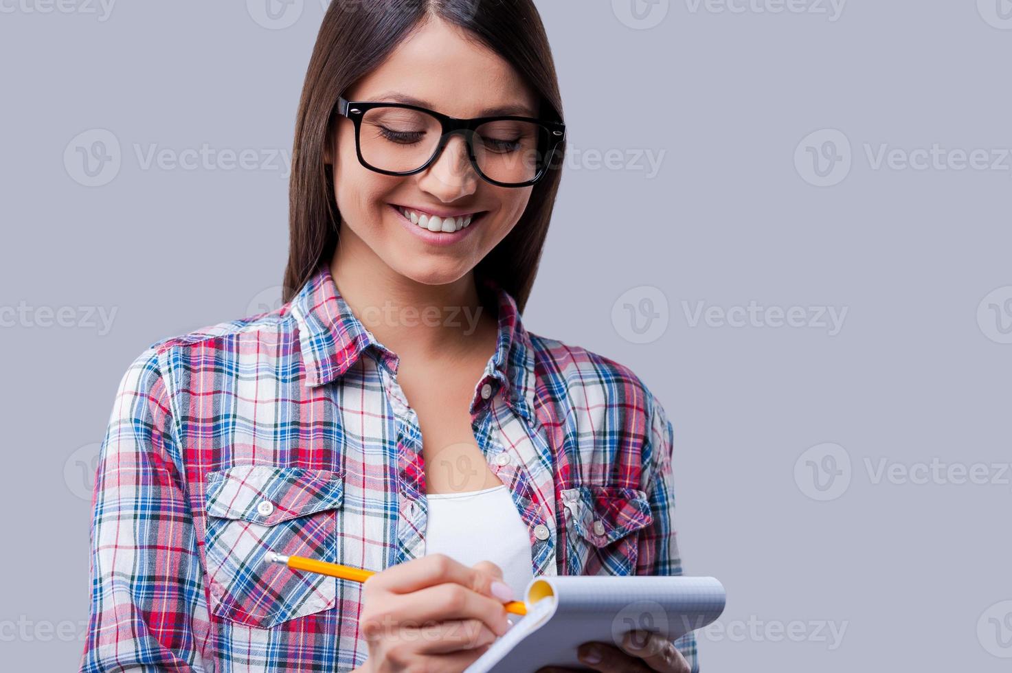 Making notes. Happy young women writing in notebook by a pencil while standing against grey background photo