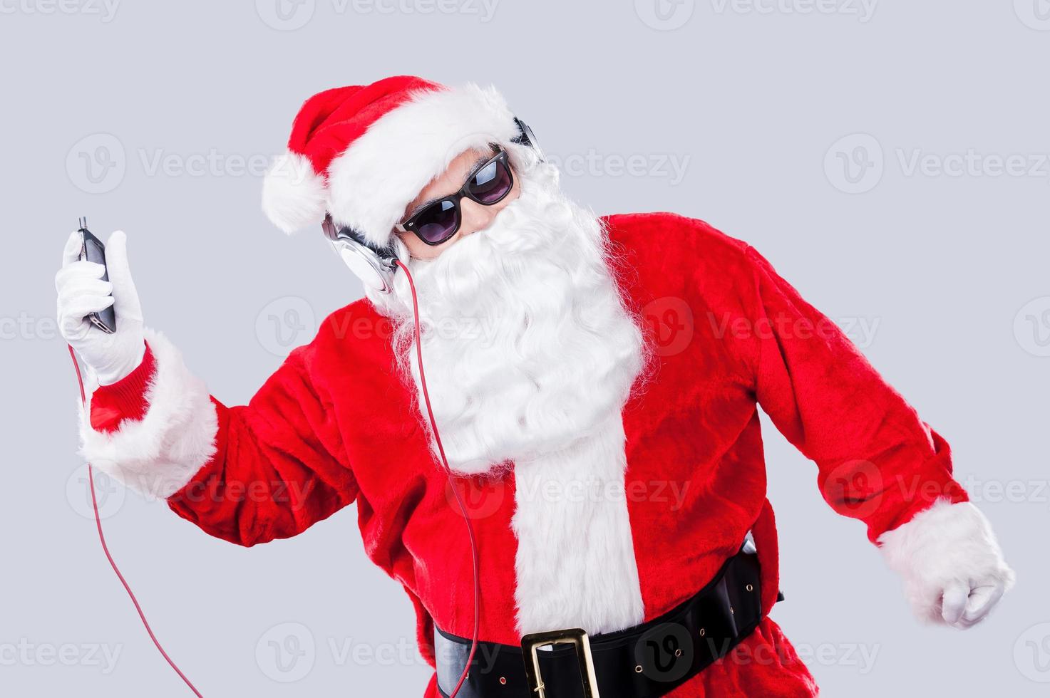 diversión navideña. santa claus con gafas de sol y auriculares escuchando un reproductor de mp3 y bailando mientras se enfrenta a un fondo gris foto