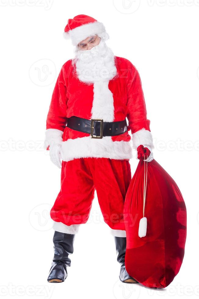 Papá Noel cansado. La longitud completa de Papá Noel cansado que lleva un saco con regalos mientras está de pie contra el fondo blanco. foto