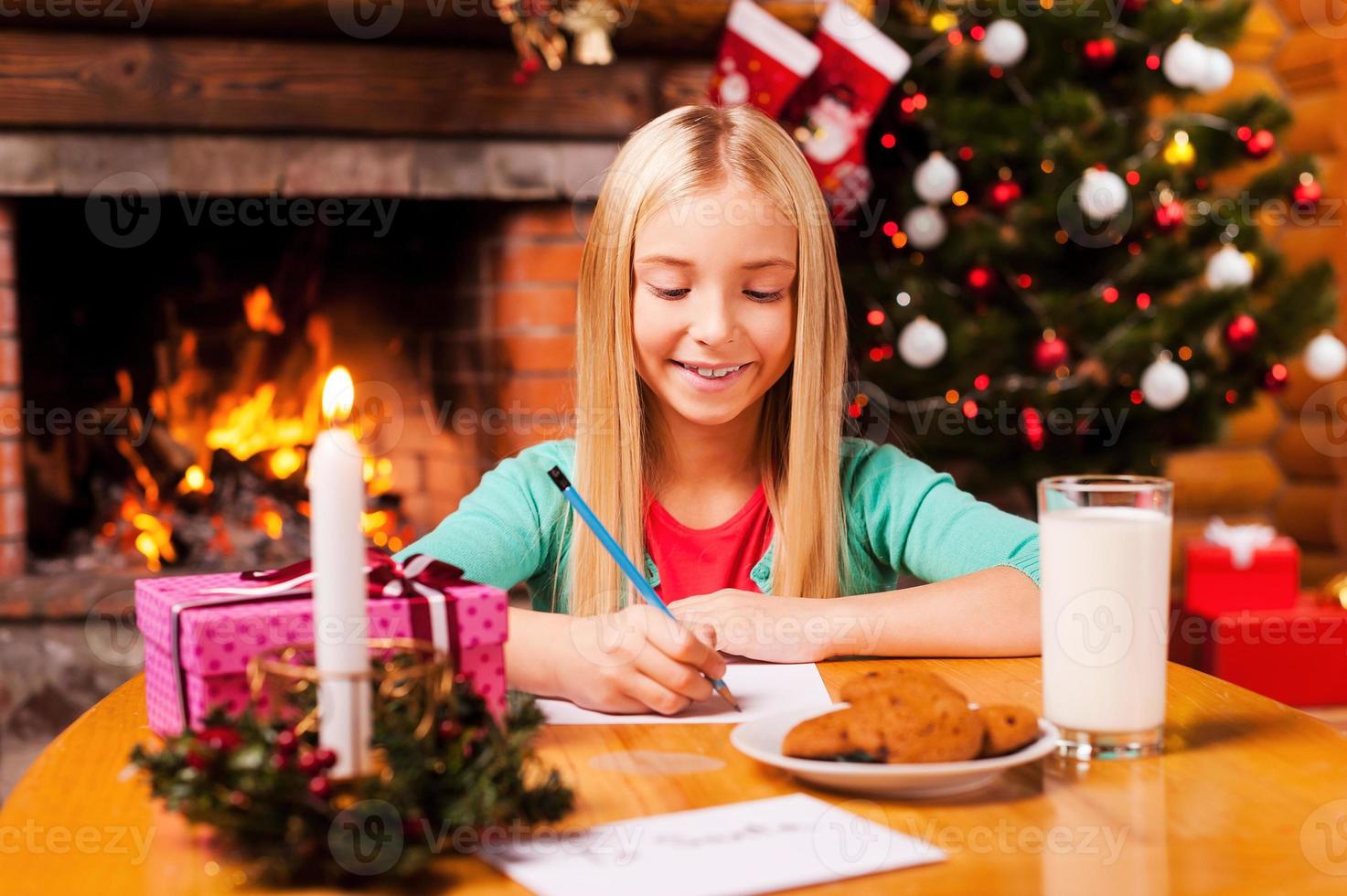 Writing a letter to Santa. Cute little girl writing a letter to Santa Claus while sitting at home with Christmas tree and fireplace in the background photo