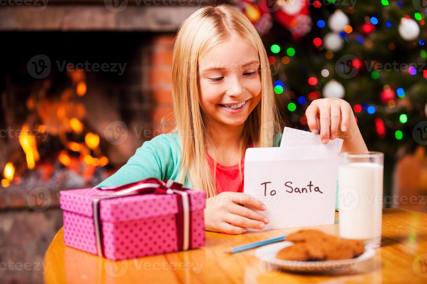 Wish list to Santa. Cute little girl putting a letter to Santa into the envelope while sitting at home with Christmas tree and fireplace in the background photo