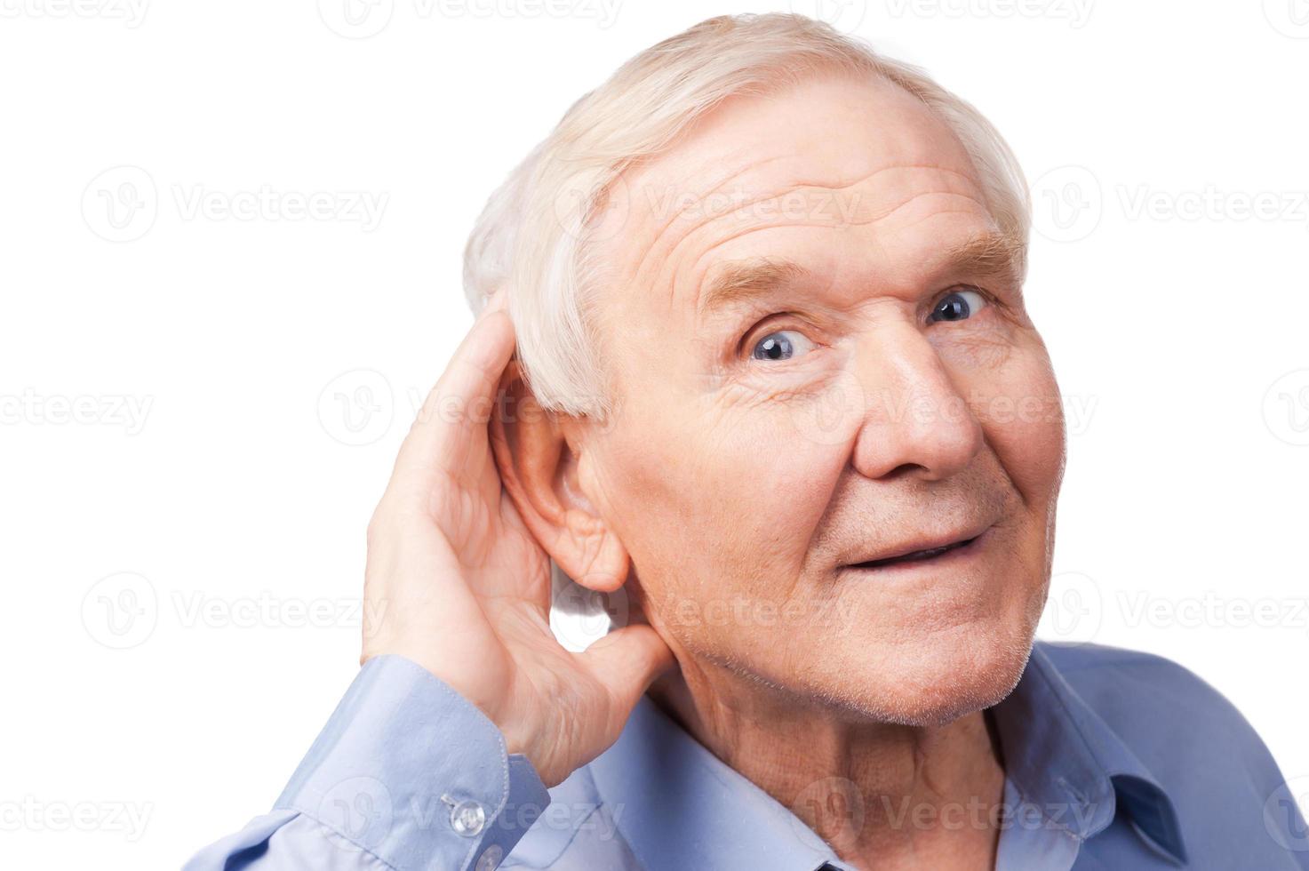 What did you say Handsome senior man holding hand near his ear and smiling while standing against white background photo