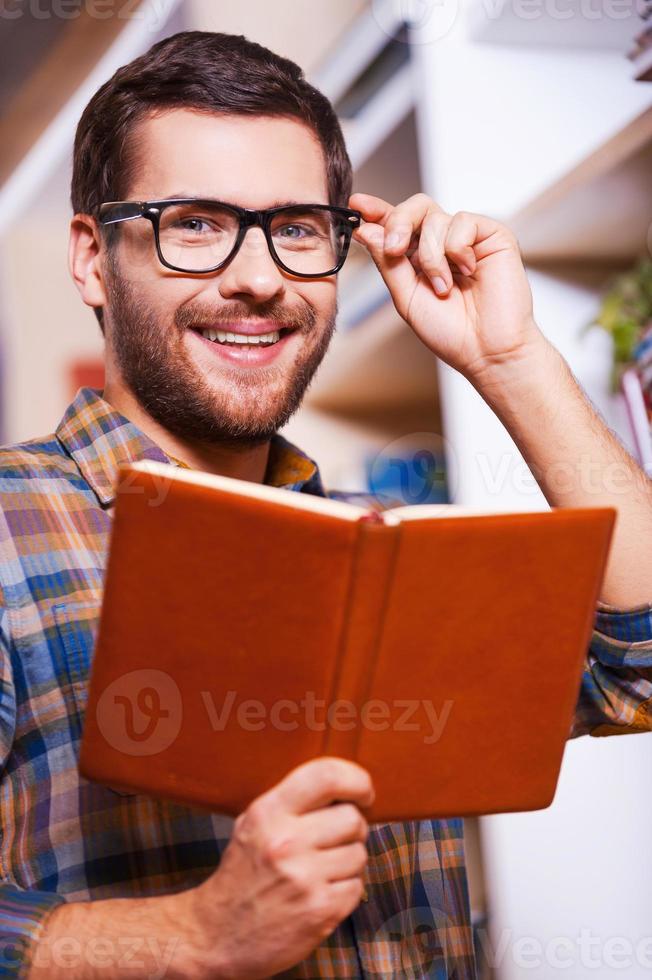 Handsome bookworm. Handsome young man holding book and smiling while standing in front of the bookshelf photo
