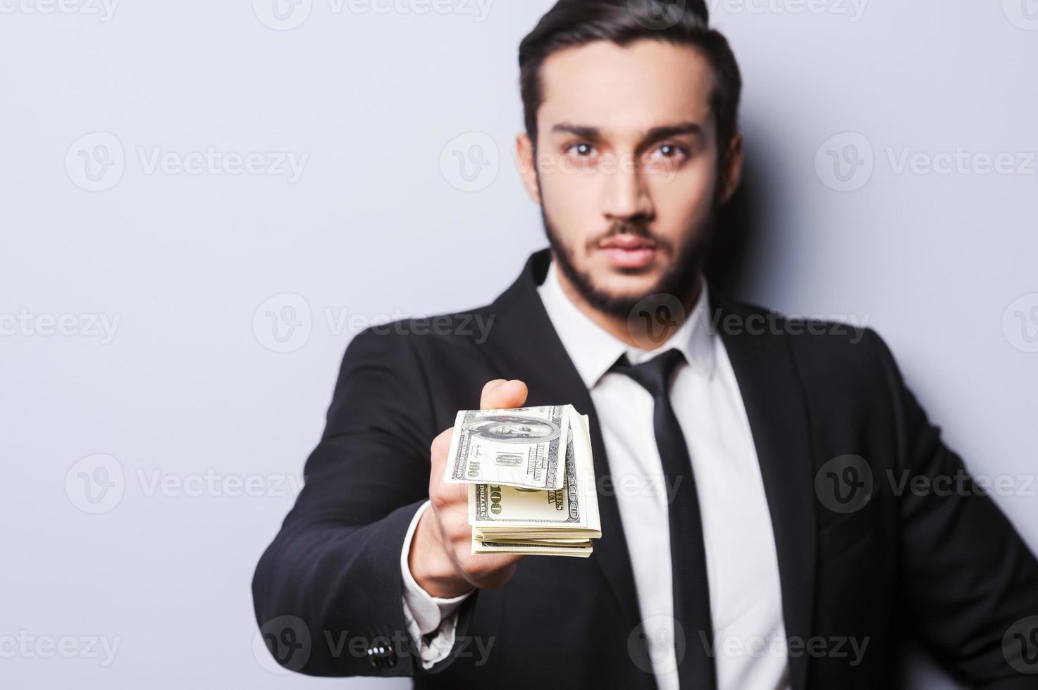 Wealthy handsome. Close-up of young man in formalwear stretching out money while standing against grey background photo