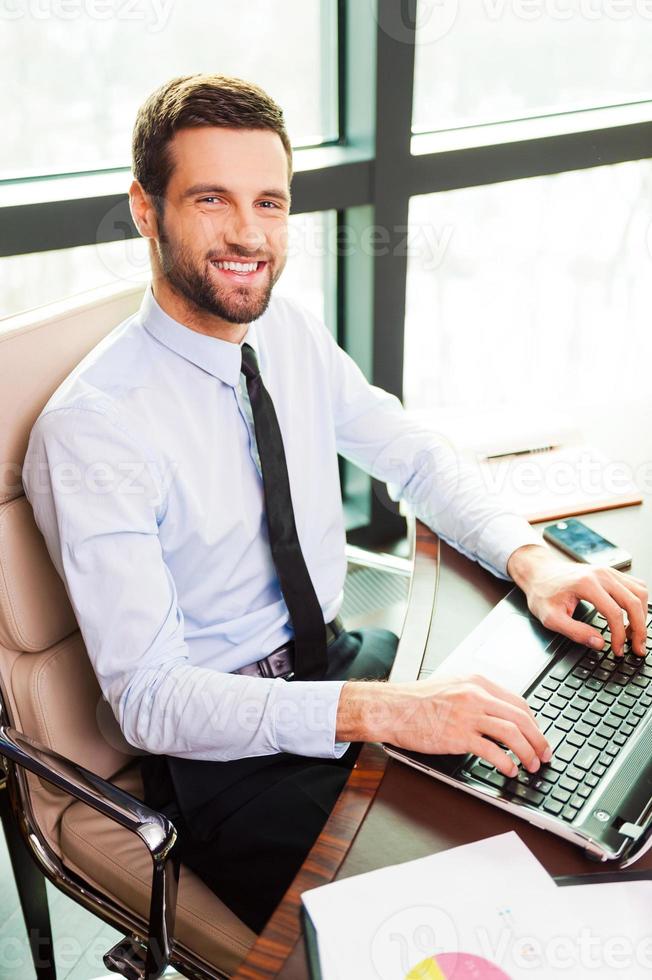 Confident businessman. Top view of handsome young man in shirt and tie looking at camera and smiling while sitting at his working place photo