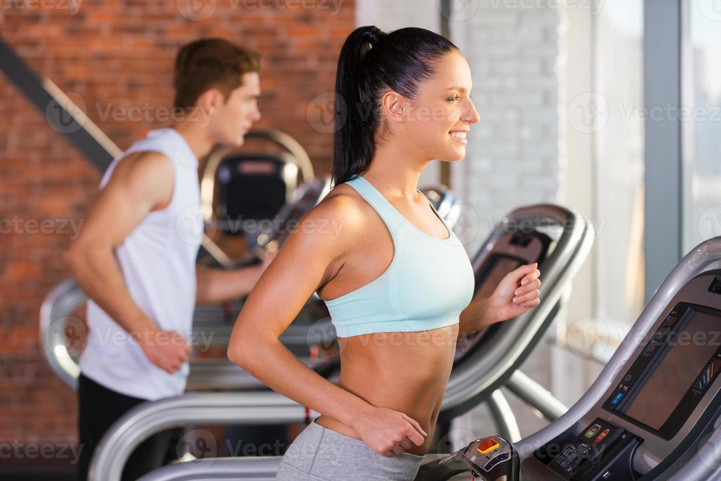 Cardio training. Side view of beautiful young woman running on a treadmill and smiling with man in the background photo