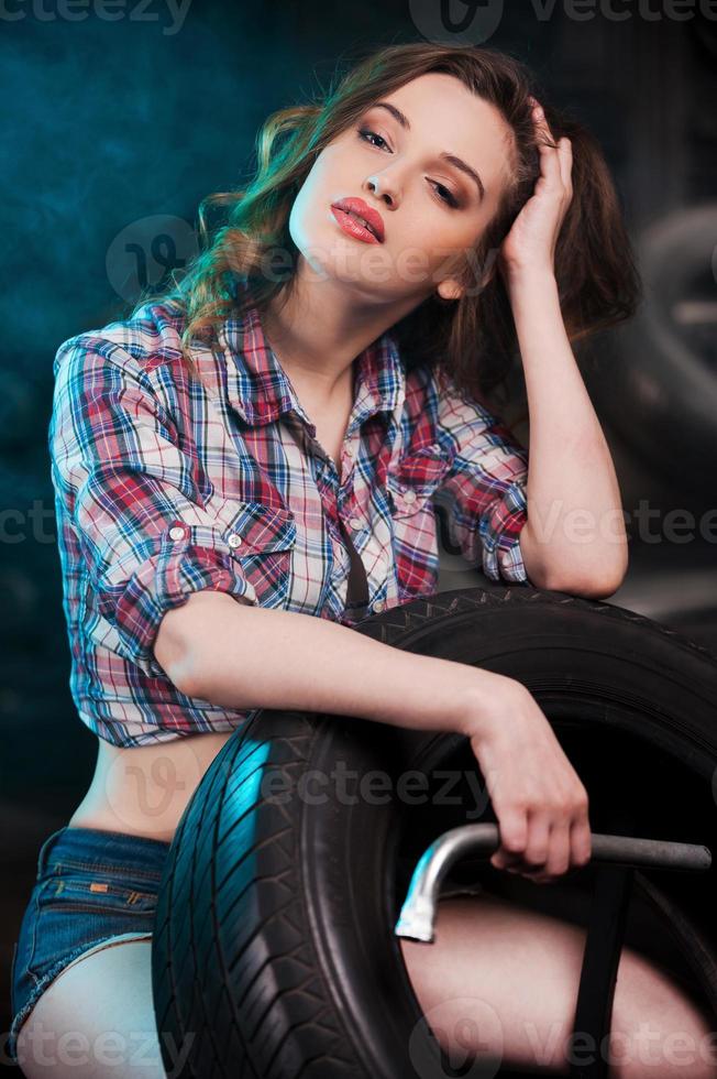 Beautiful mechanic. Beautiful young woman leaning at the car tire and looking at camera while sitting in auto repair shop photo