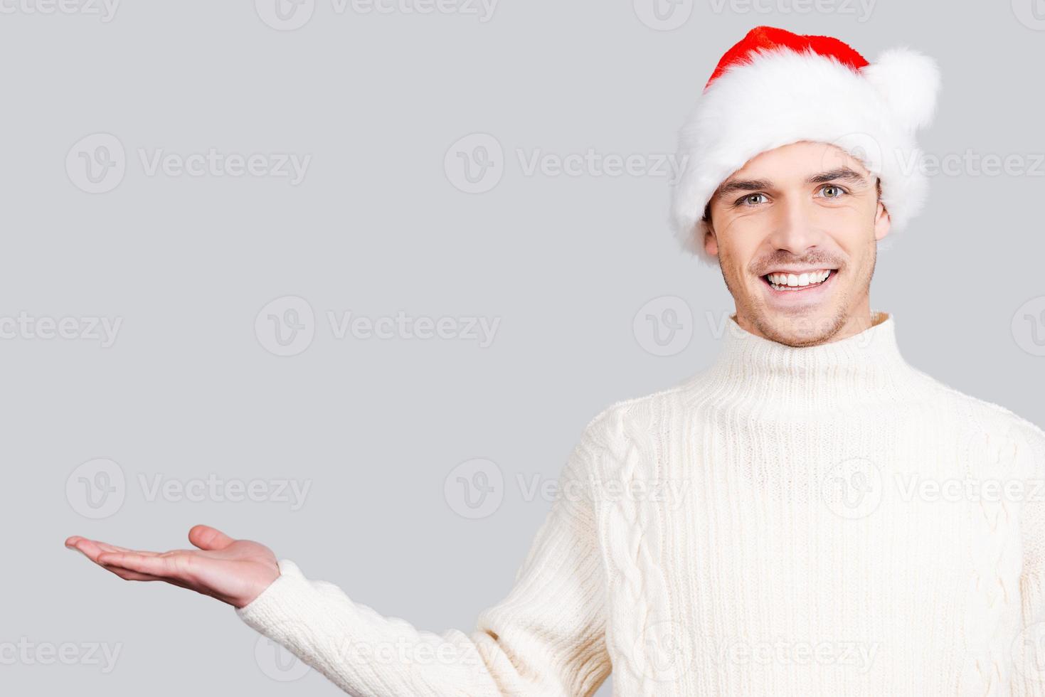 Copy space in my hand. Handsome young man in Santa hat holding copy space and smiling while standing against grey background photo