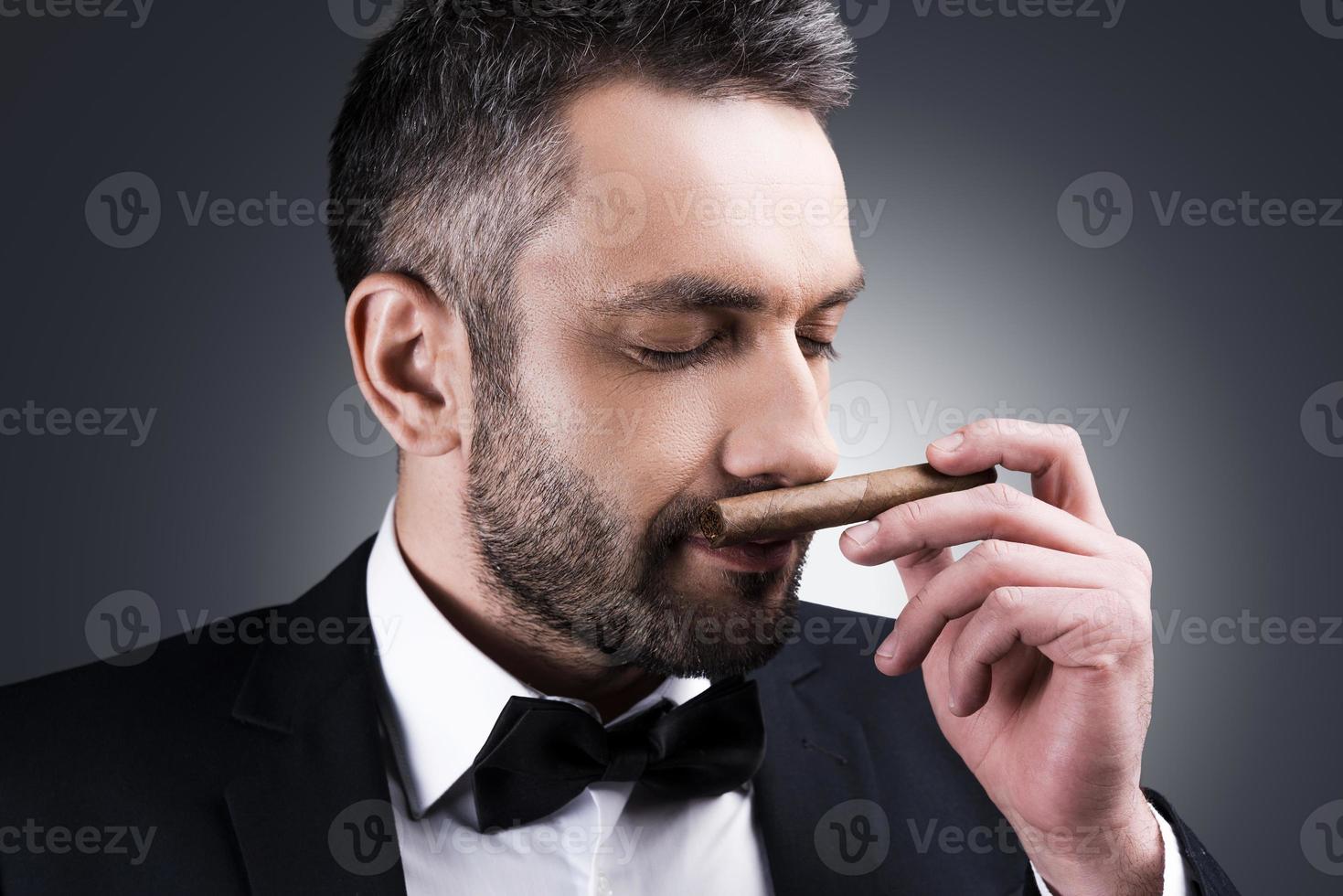 gran retrato de cigarro de un apuesto hombre maduro con ropa formal que huele a cigarro y mantiene los ojos cerrados mientras se enfrenta a un fondo gris foto