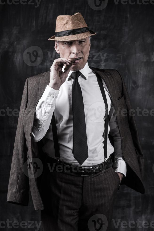 Powerful boss. Serious senior man in hat and suspenders smoking cigar and looking at you while standing against dark background photo