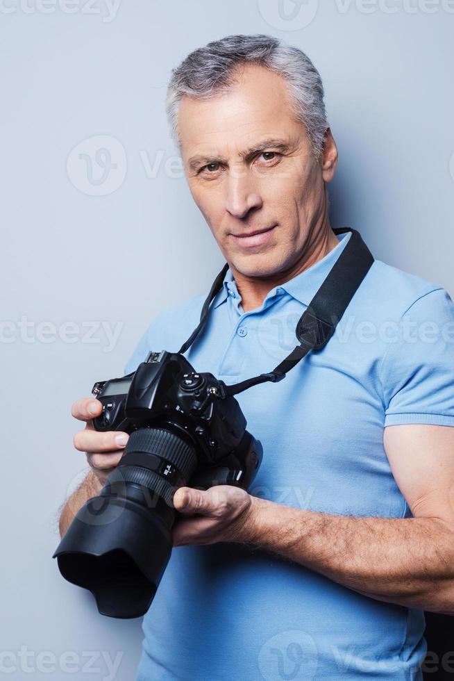 Favorite activity brings maximum pleasure. Portrait of confident senior man in T-shirt holding camera while standing against grey background photo