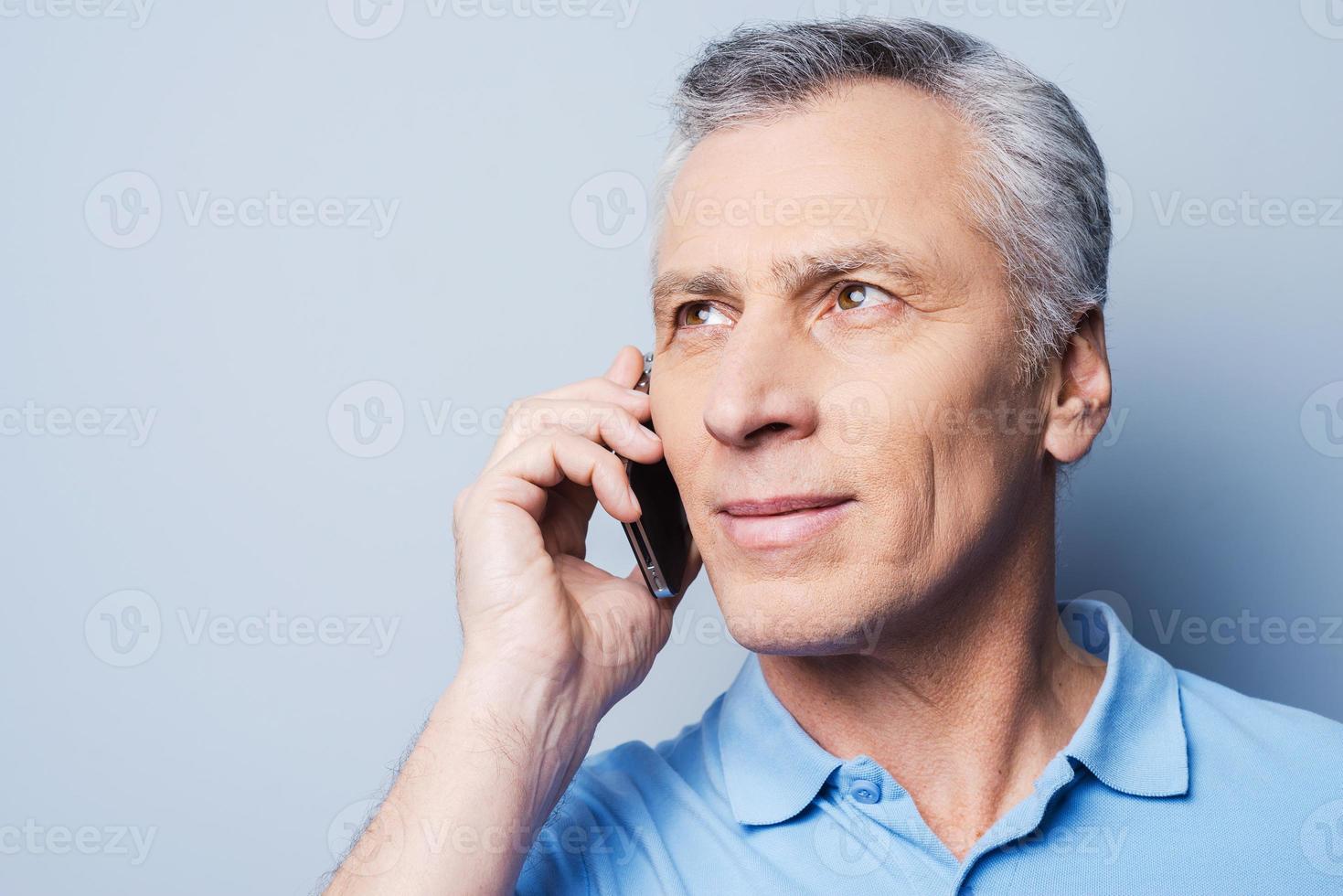Good talk. Portrait of confident senior man in shirt talking on the mobile phone and smiling while standing against grey background photo
