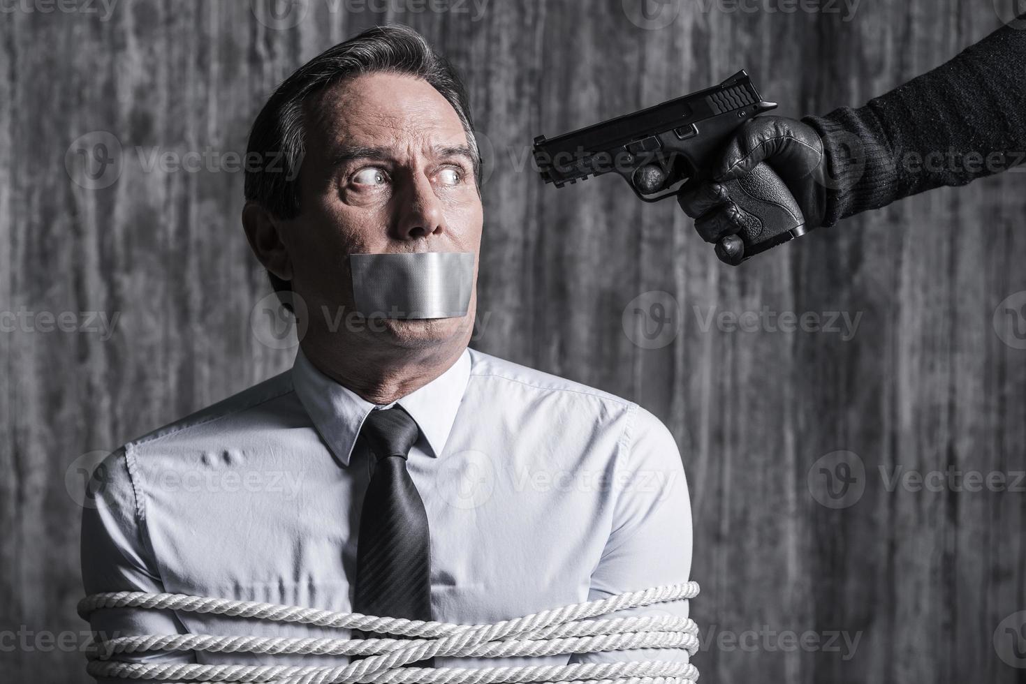 Please no. Tied up businessman with adhesive tape on his mouth sitting in front of the dirty wall while someone aiming his head photo
