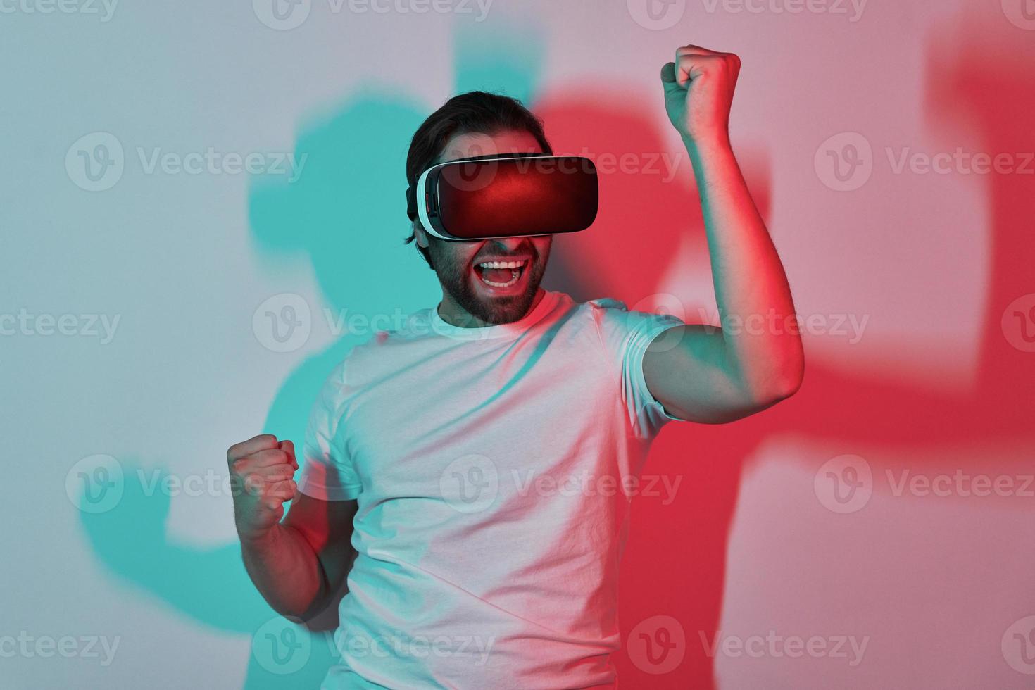 joven feliz con anteojos de realidad virtual gesticulando con sombras coloridas en el fondo foto