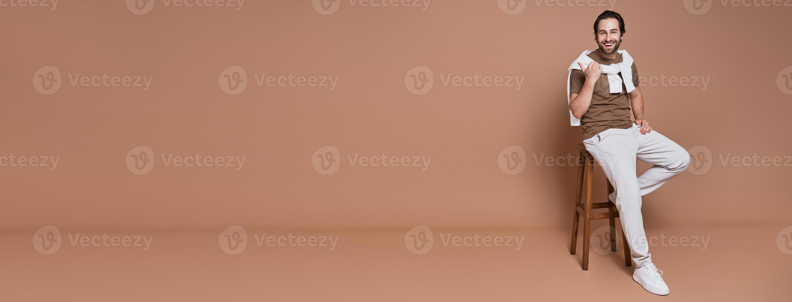 Happy young man pointing away and smiling while sitting against brown background photo