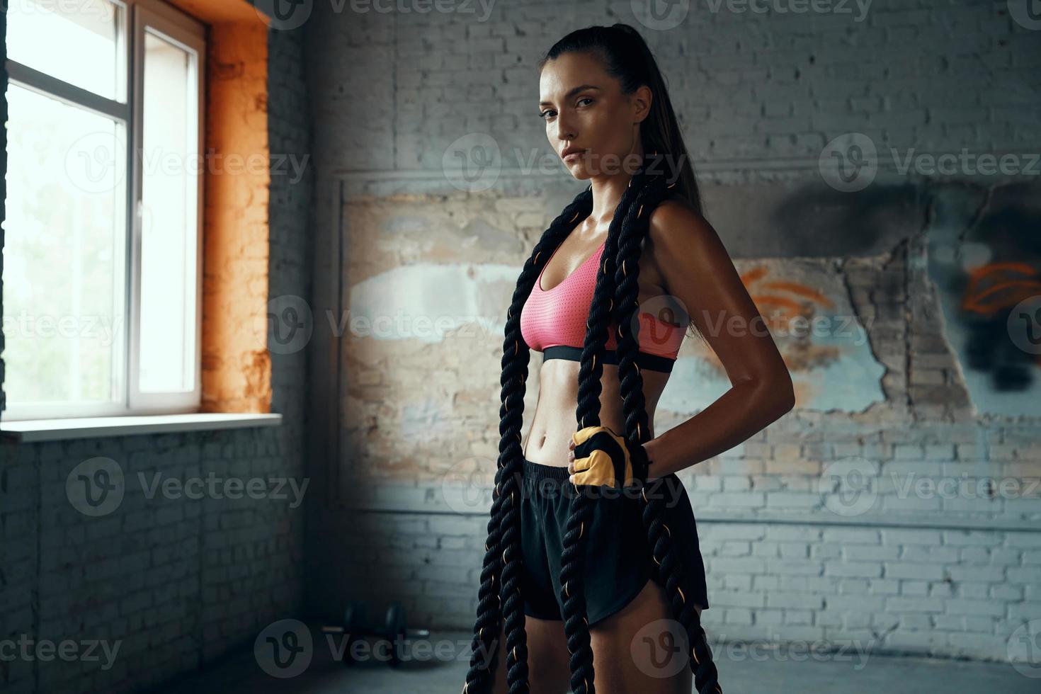 Beautiful young woman carrying battle rope and looking at camera while standing in gym photo