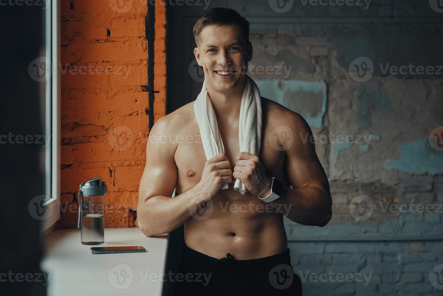 Handsome muscular man carrying towel on shoulders and smiling while standing in gym photo