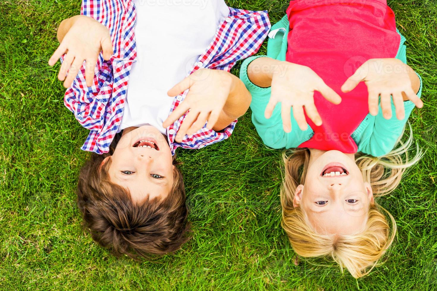 We love summer Top view of two cute little children raising their hands up and smiling while lying on the green grass together photo