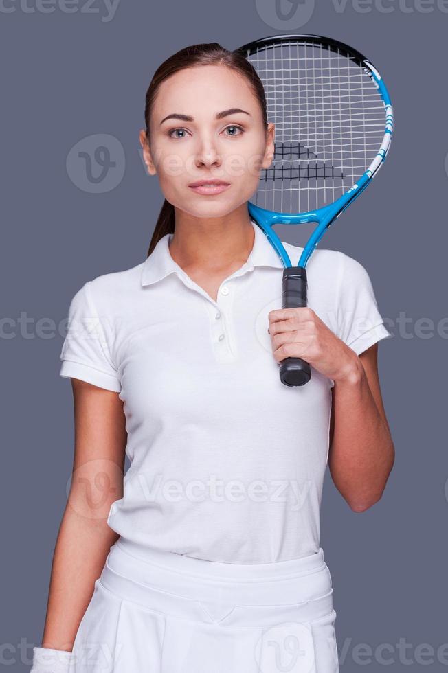 Serious and sporty.  Beautiful young women in sports clothes holding tennis racket on her shoulder and looking at camera while standing against grey background photo