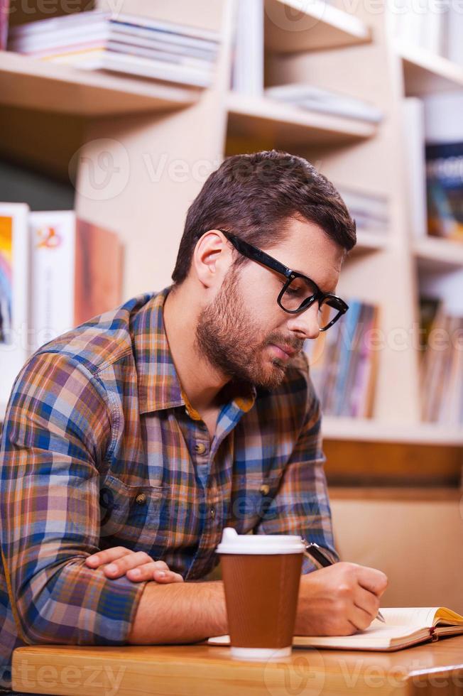 Spending time in library. Confident young man writing something in note pad while sitting at the desk in library photo