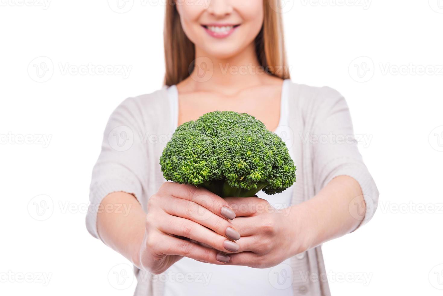 Giving you a pit of vitamins. Cropped image of beautiful young woman stretching out broccoli and smiling while standing against white background photo