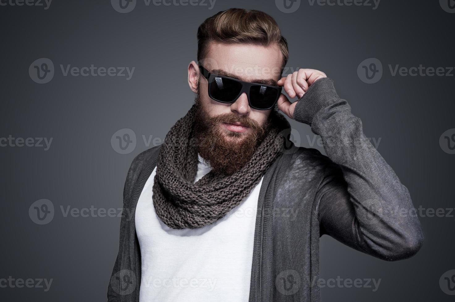 Looking cool and trendy. Handsome young bearded man adjusting his sunglasses and looking at camera while standing against grey background photo