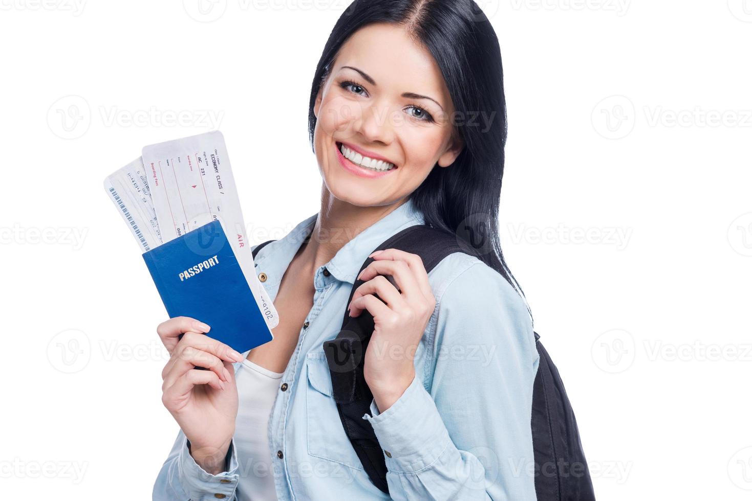 To another country for the new emotions. Beautiful young smiling woman holding tickets and passport while standing against white background photo