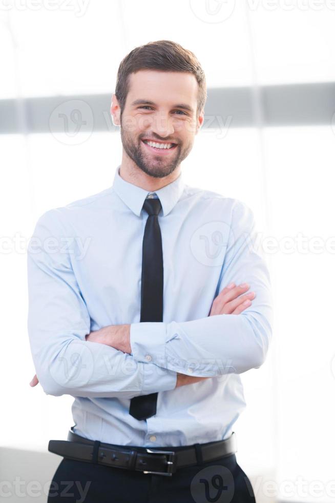 Confident business expert. Confident young businessman keeping arms crossed and smiling while standing indoors photo