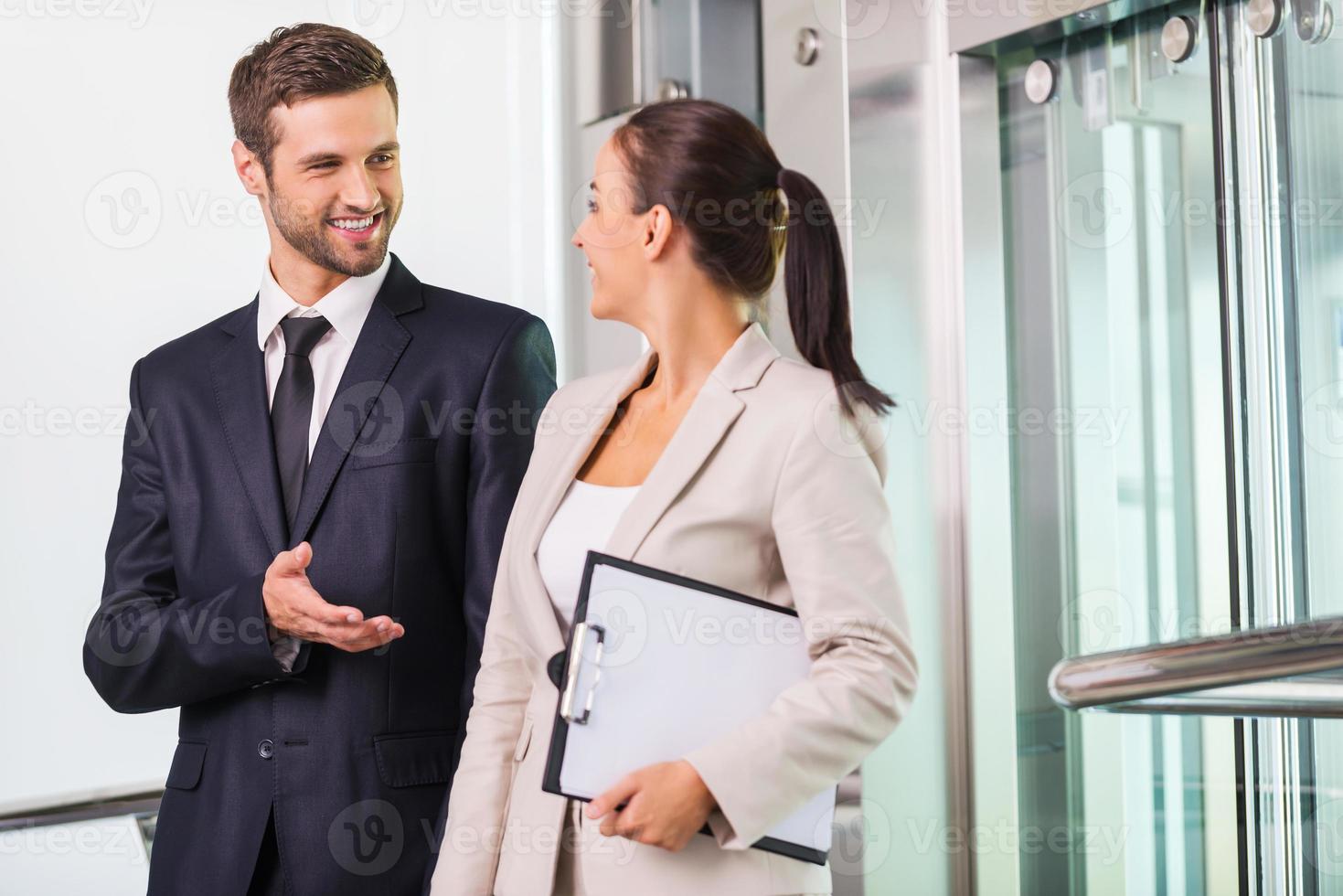 Discussing project with colleague. Two cheerful business people discussing something and smiling while getting out from elevator photo