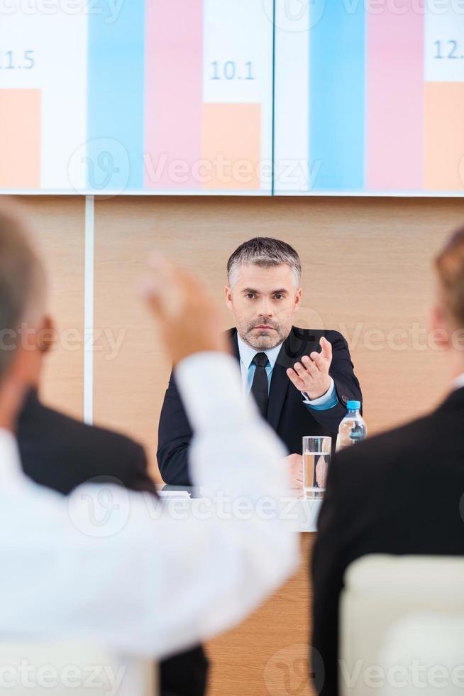 Everyone is welcome for discussion. Confident mature man in formalwear giving a word to someone from audience while sitting in conference hall photo