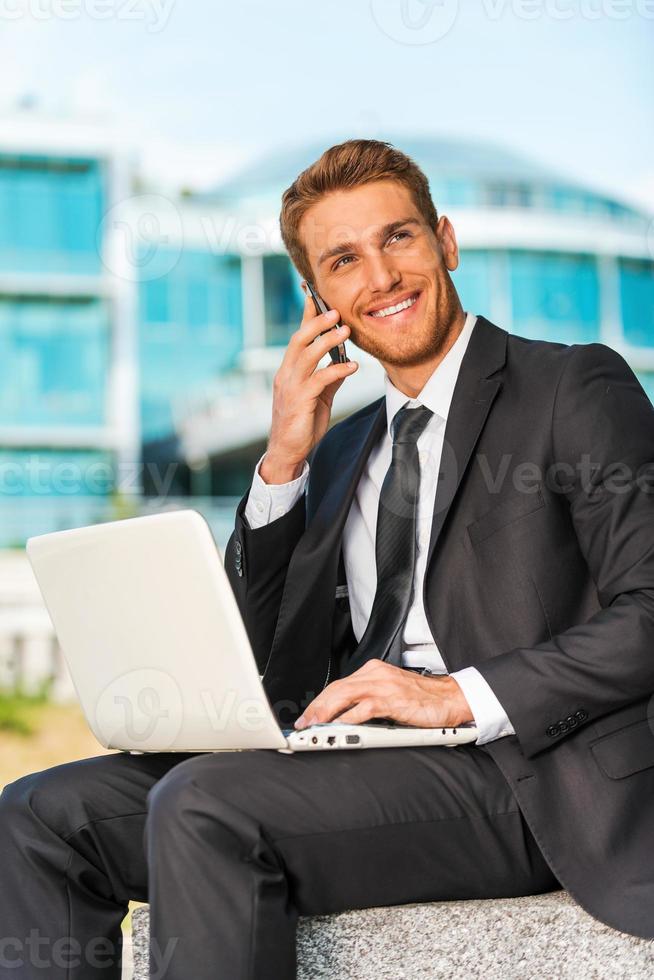 Businessman working outdoors. Handsome young man in formalwear working on laptop and talking on the mobile phone while sitting outdoors and against building structure photo