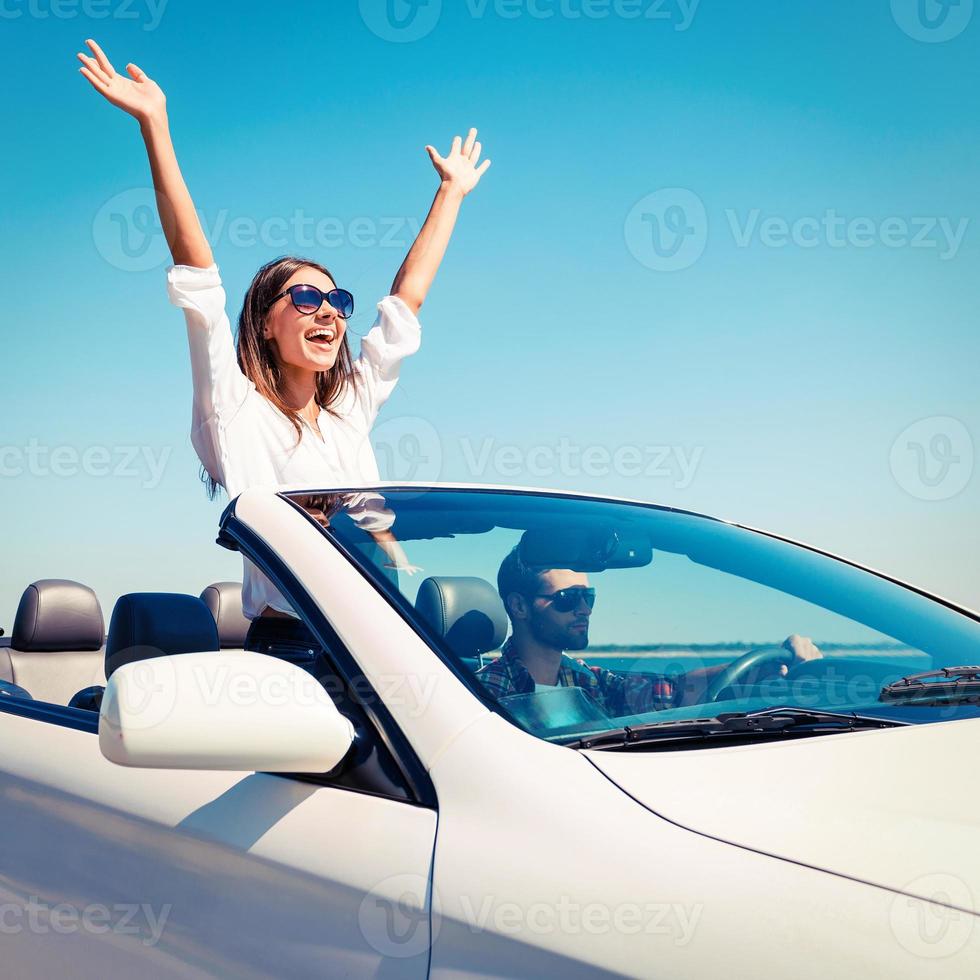 Couple in convertible. Happy young couple enjoying road trip in their convertible while woman raising arms and smiling photo