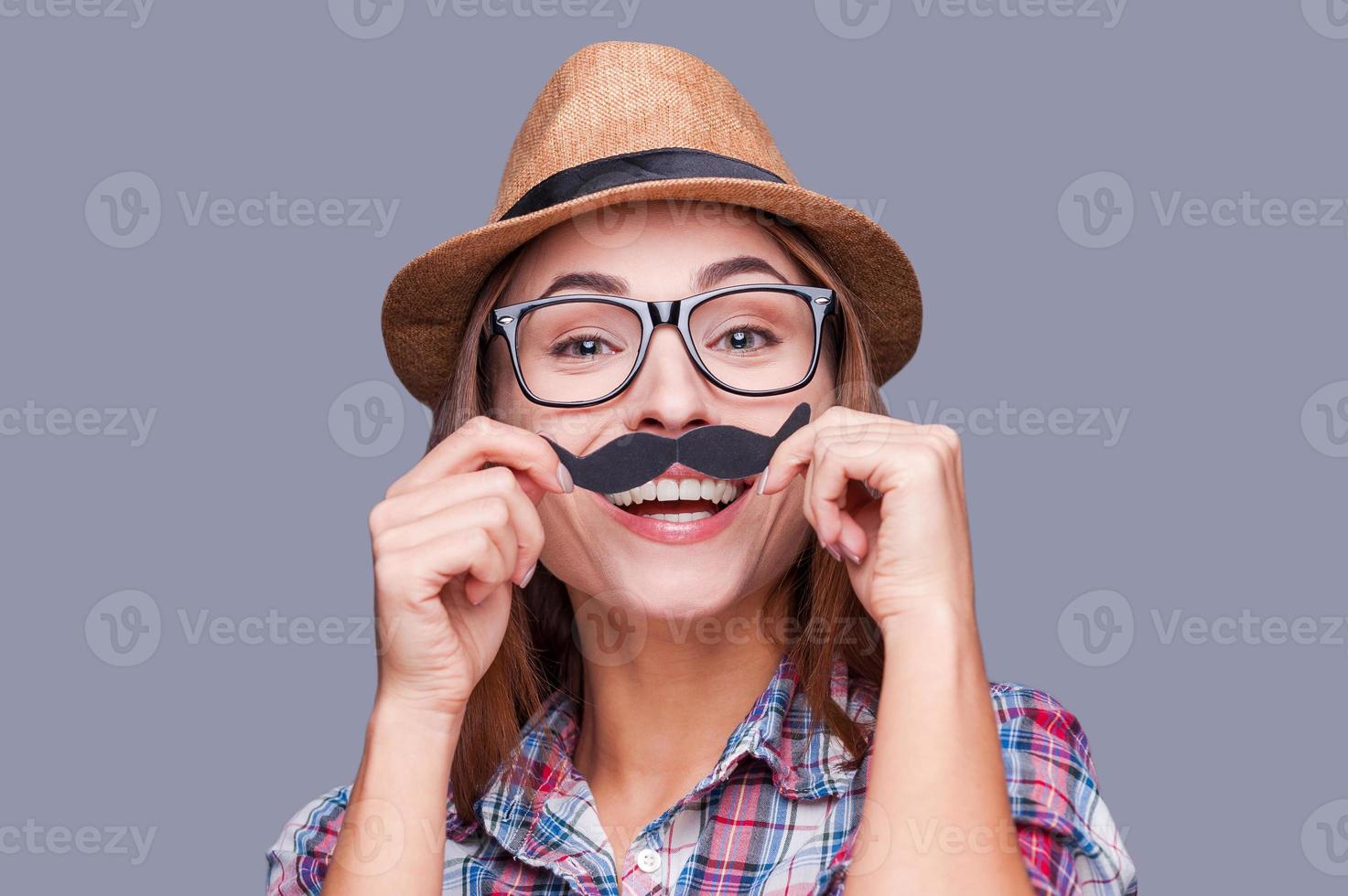 Having fun with face mustache. Cheerful young woman in funky hat holding fake mustache on her face and looking at camera while standing against grey background photo