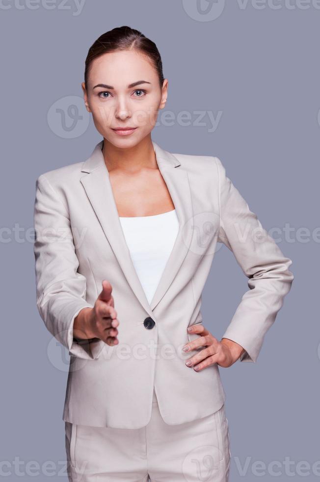 Welcome on board Confident young businesswoman stretching out hand for shaking while standing against grey background photo