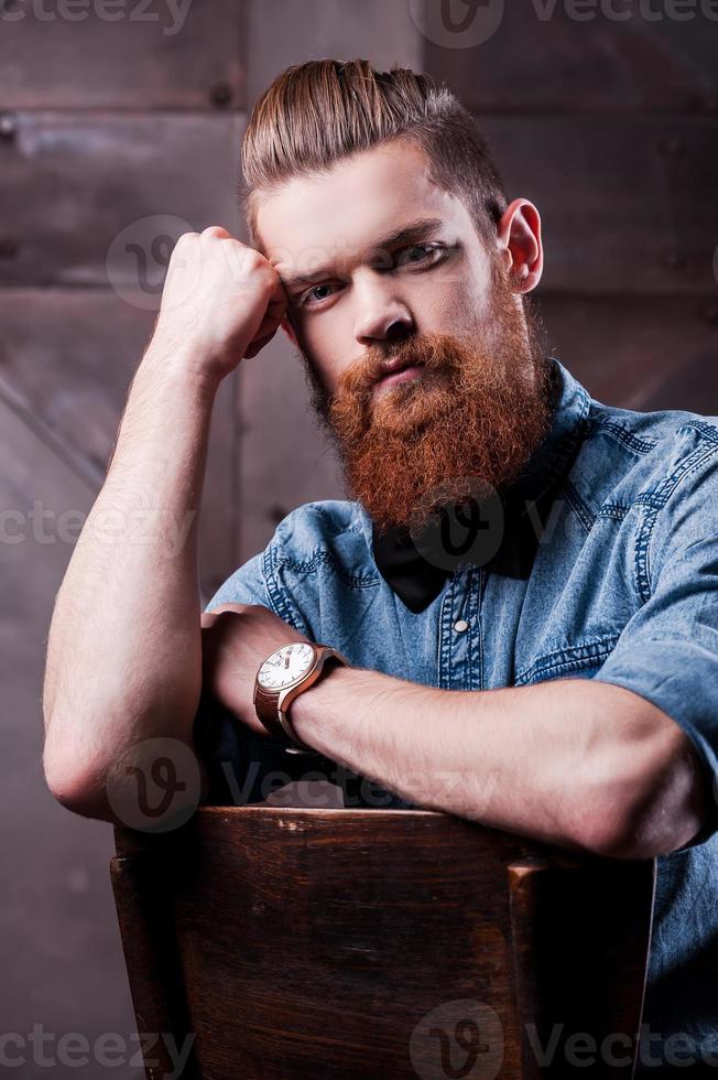 Stylish handsome. Handsome young bearded man holding head in hand and looking at camera while sitting on chair photo