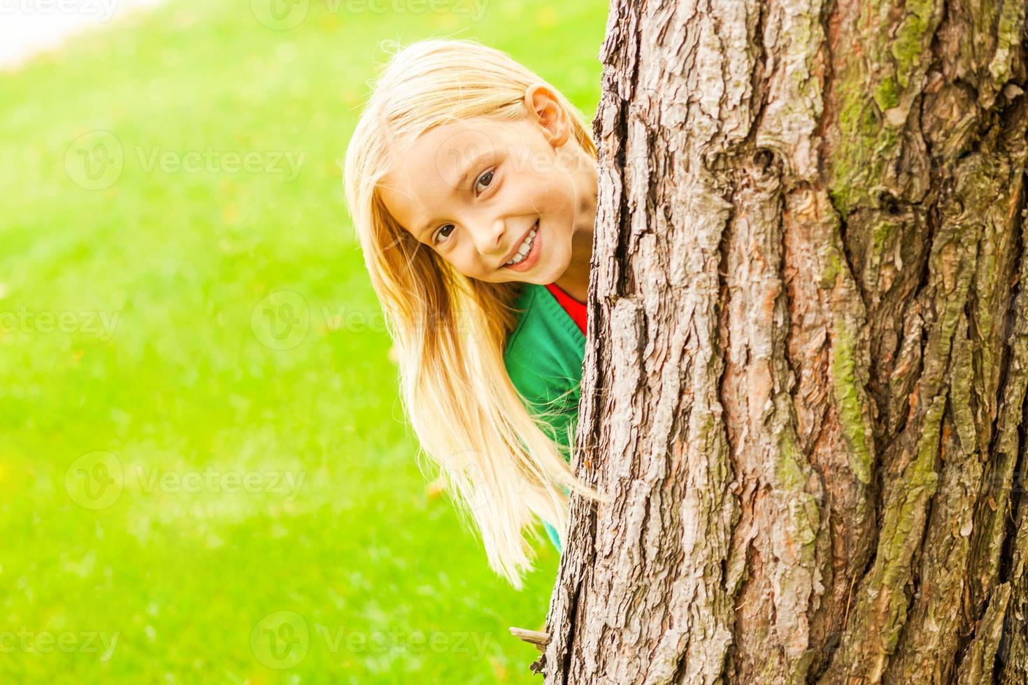 sintiéndose juguetón. linda niña mirando por el árbol y sonriendo mientras está de pie al aire libre foto