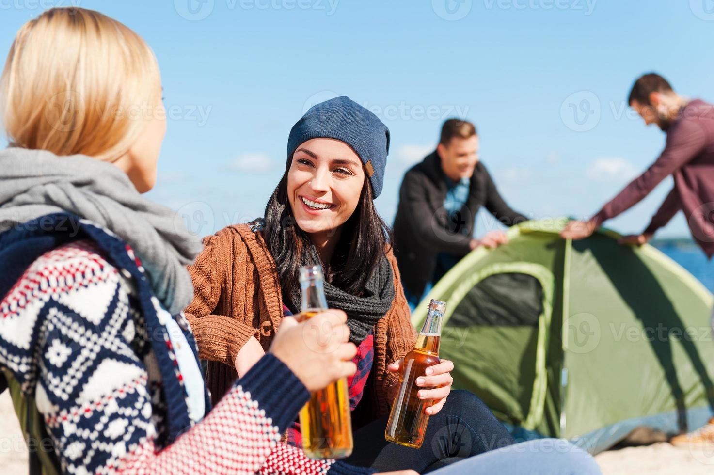 Taking time to have a rest. Two beautiful young women talking to each other and smiling while holding bottles with beer with two men setting up tent in the background photo