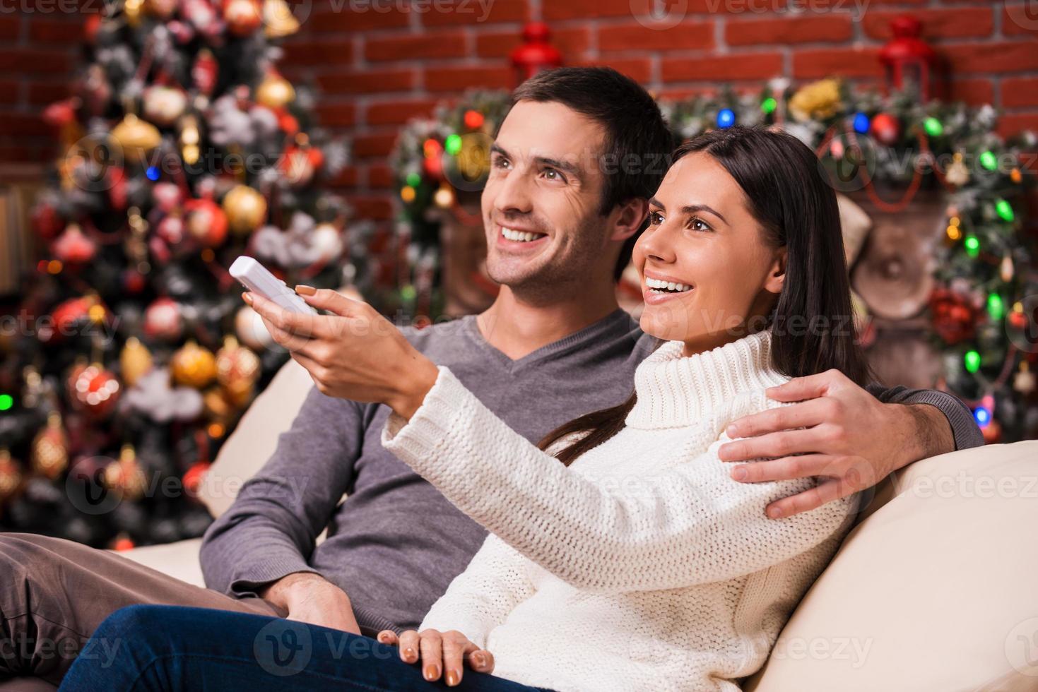 viendo programas navideños juntos. vista lateral de una hermosa pareja joven y amorosa que se une y sonríe mientras ve la televisión junto con el árbol de Navidad en el fondo foto