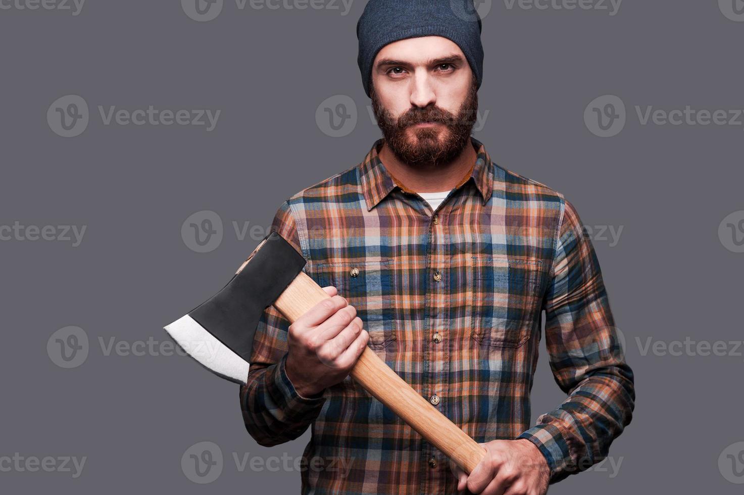 Serious lumberjack. Confident young bearded man holding a big axe and looking at camera while standing against grey background photo