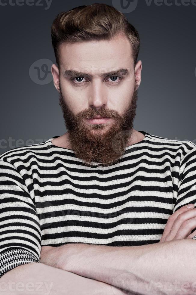 confianza y masculinidad. un joven barbudo confiado vestido a rayas con los brazos cruzados y mirando a la cámara mientras se enfrenta a un fondo gris foto