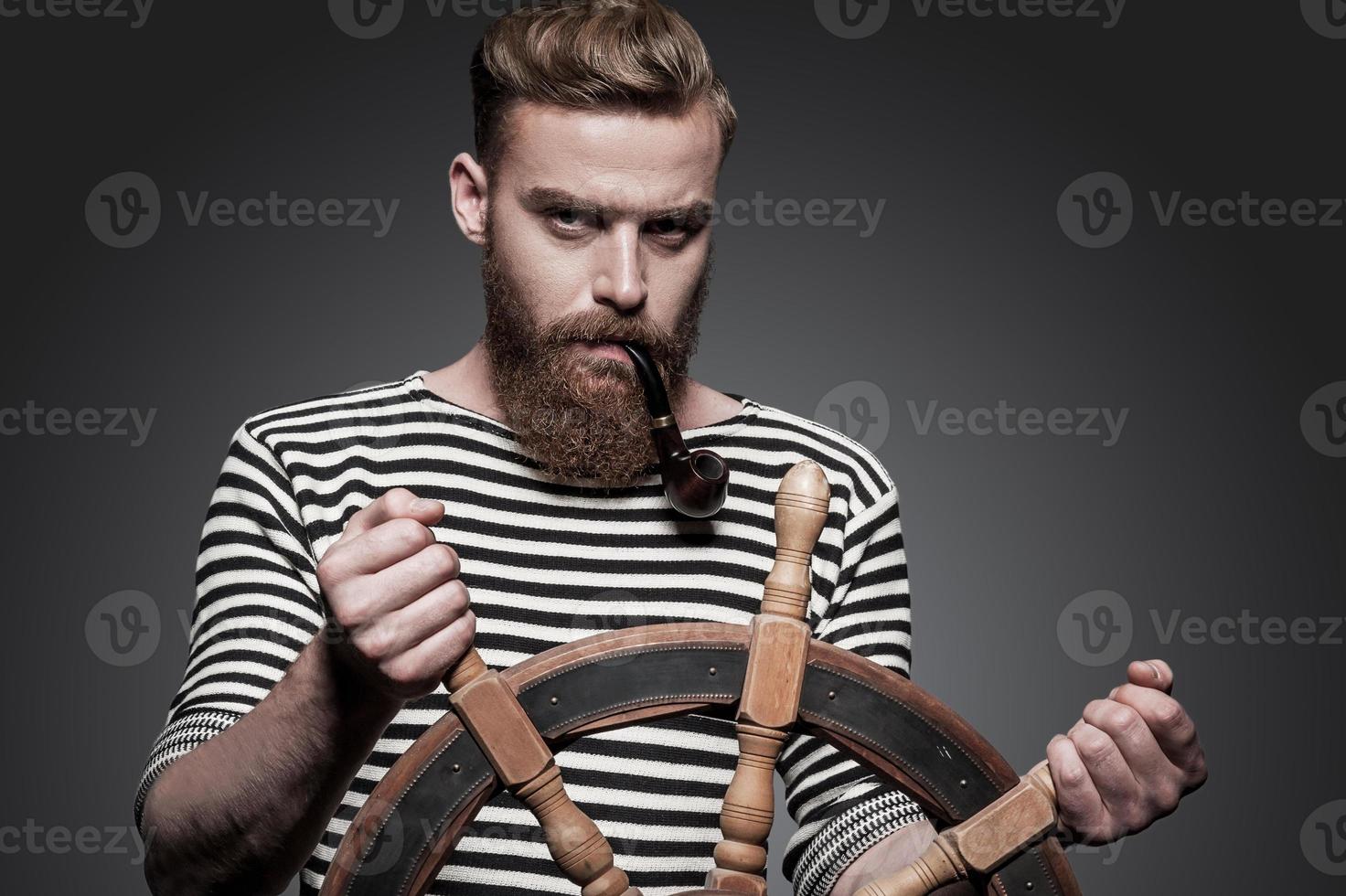 Finding the right way. Confident young bearded sailor steering a wheel while standing against grey background photo