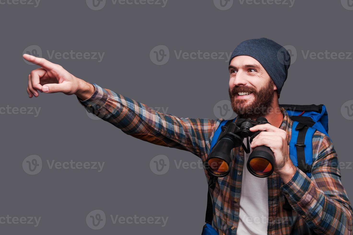 That is amazing Handsome young bearded man with backpack holding binoculars and pointing away with smile while standing against grey background photo