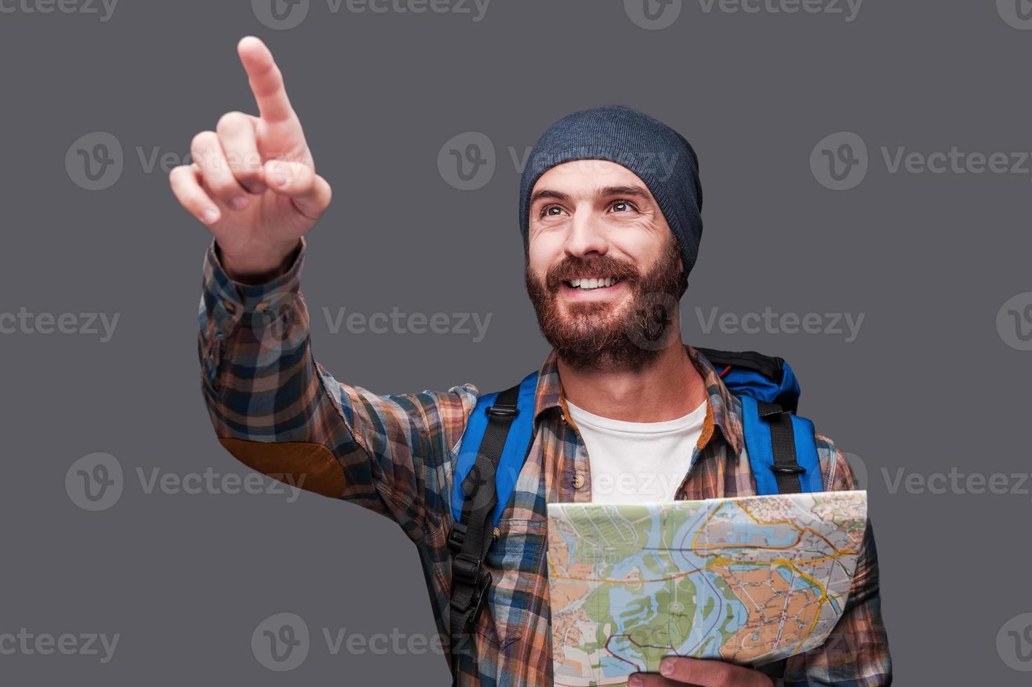 sé la dirección correcta feliz joven barbudo con mochila sosteniendo un mapa y señalando mientras está de pie contra un fondo gris foto