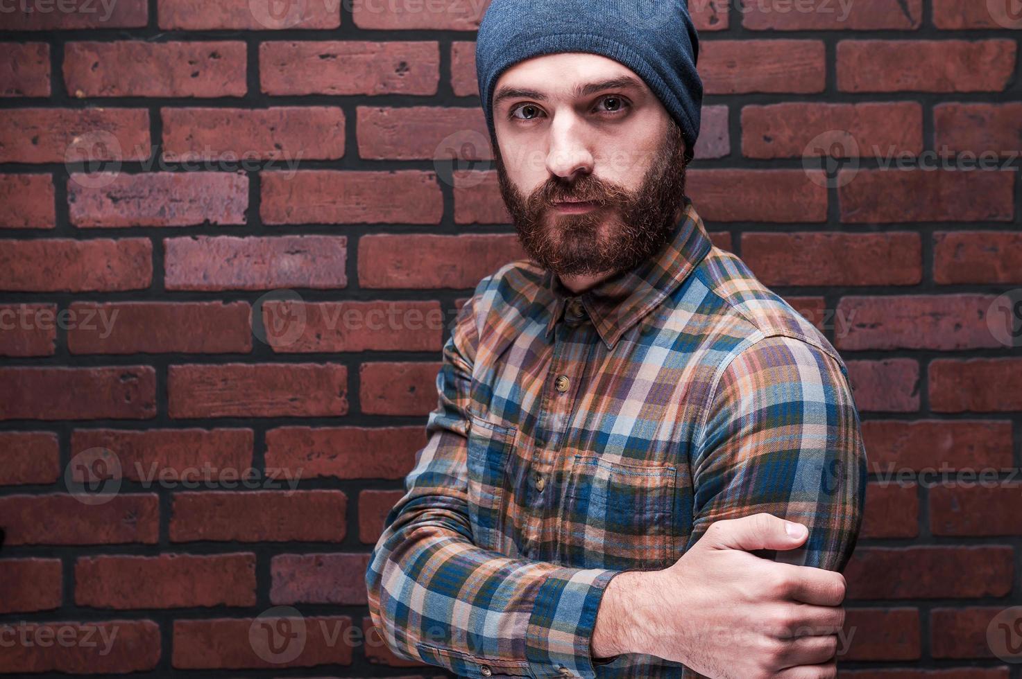 Bearded handsome. Handsome young bearded man looking at camera while standing against brick wall photo