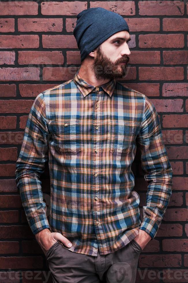 Handsome man in style. Handsome young bearded man holding hands in pockets and looking away while standing against brick wall photo